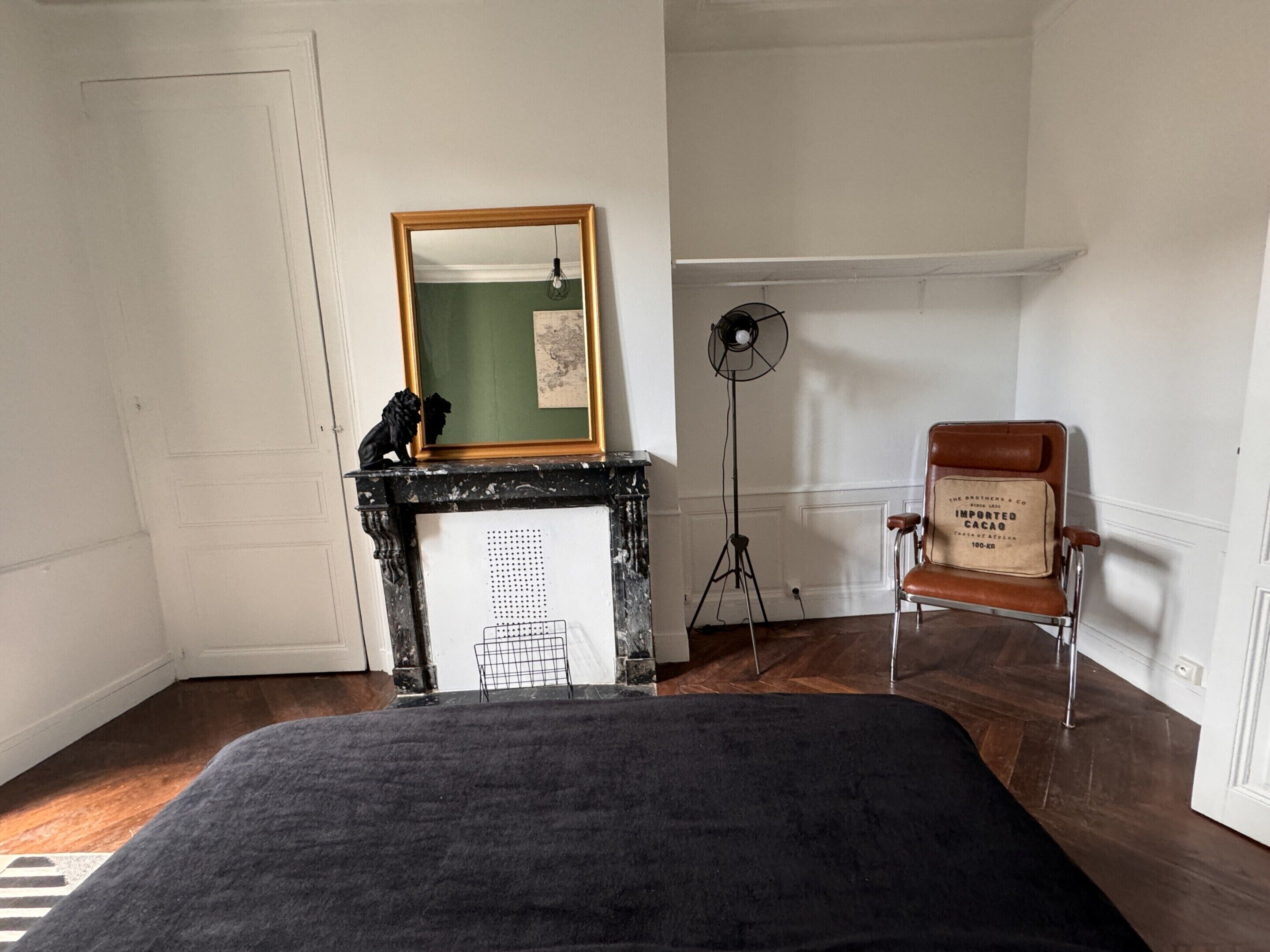 A room with a bed, a fireplace with a mirror above it, a floor lamp, and a vintage chair.