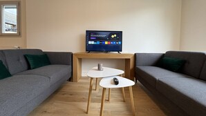A modern living room with a flat-screen TV on a wooden stand, two grey sofas with green cushions, and a small white table with a remote control. - Le Corner - Ville - 8 Pers (Le Mans)