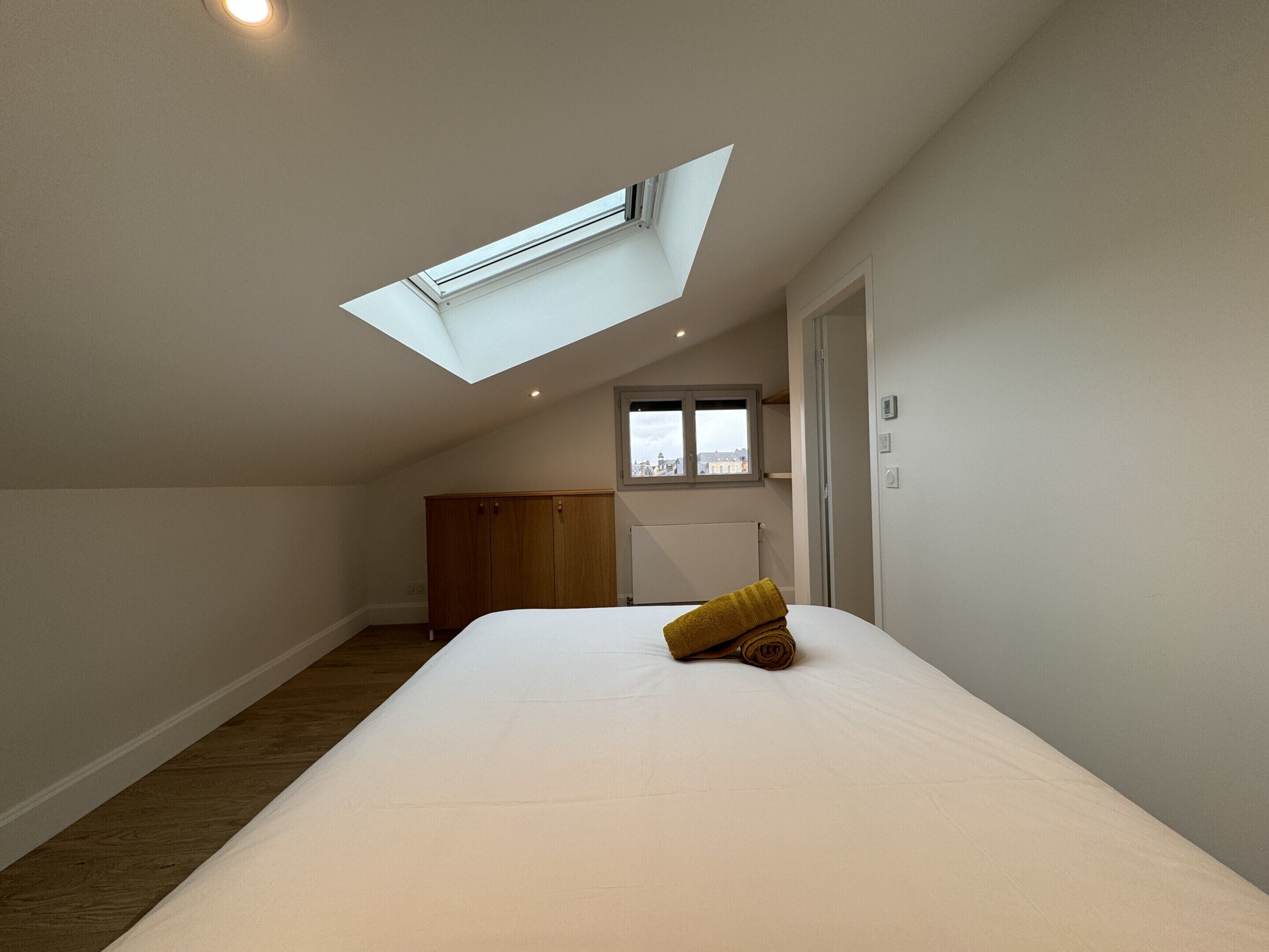A bedroom with a skylight, a bed, a wardrobe, and a window with a view of buildings.