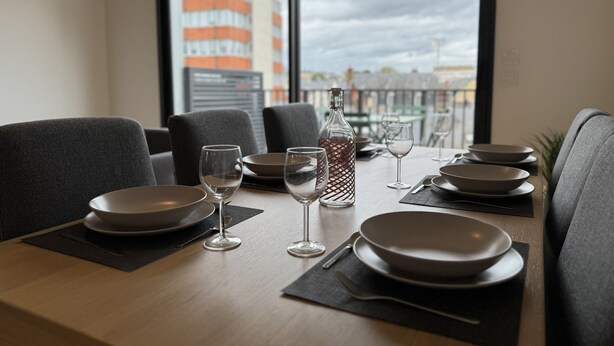 A dining table set with plates, glasses, and a bottle, with a view of buildings through a window. - Le Corner - Ville - 8 Pers (Le Mans)