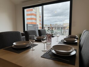 A dining table set with plates, glasses, and a bottle, with a view of buildings through a window. - Le Corner - Ville - 8 Pers (Le Mans)