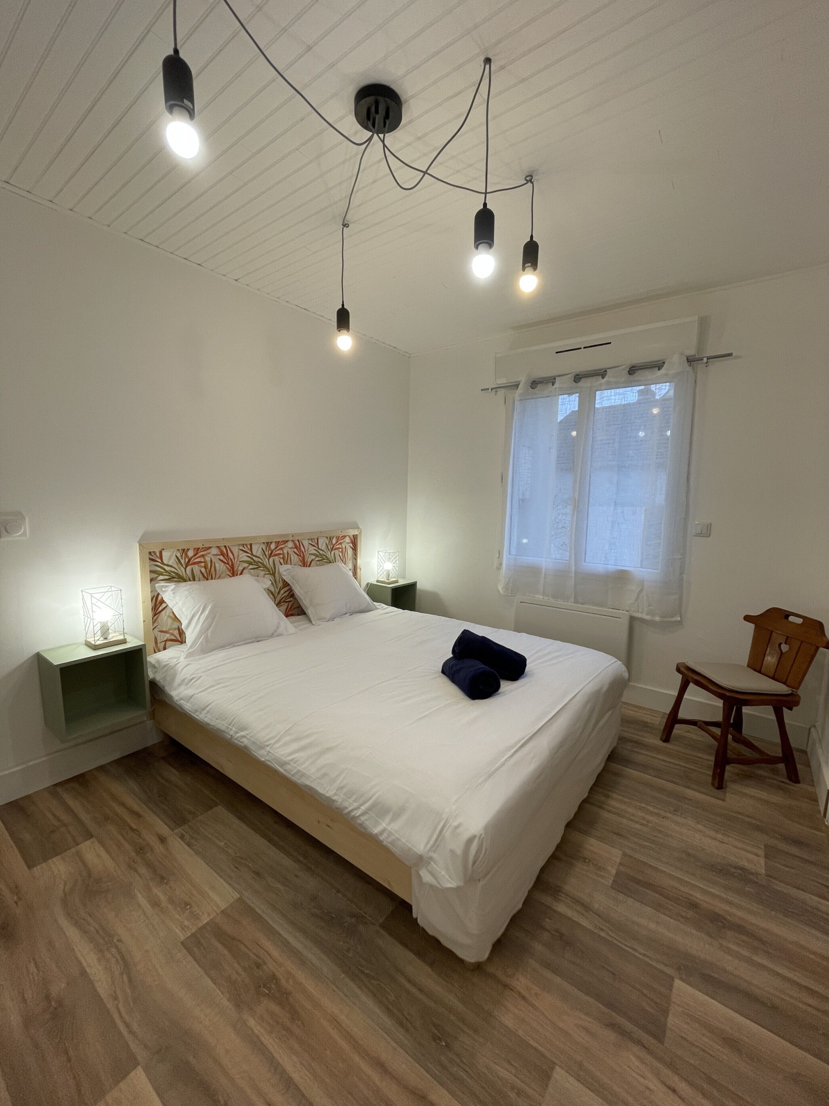 A neatly made bed with white linens, a wooden headboard, and a pair of rolled towels on the bed. There is a wooden chair and a nightstand with a lamp next to the bed. The room has wooden flooring and a window with curtains.