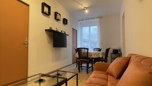 A living room with a glass-top coffee table, a sofa, a dining area with a table and chairs, and a wall-mounted clock. - 3-bedroom Apartment With Premium Amenities (Noyen-sur-Sarthe)