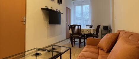 Un salon comprenant une table basse avec un plateau en verre, un canapé, un coin repas avec une table et des chaises, et une horloge fixée au mur.
