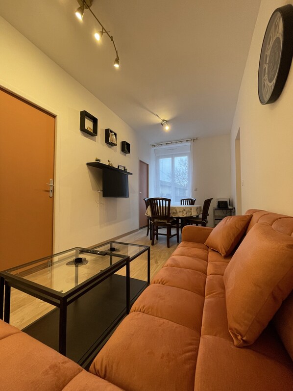 Una sala de estar con una mesa de centro de vidrio, un sofá, un comedor con mesa y sillas, y un reloj fijado a la pared.