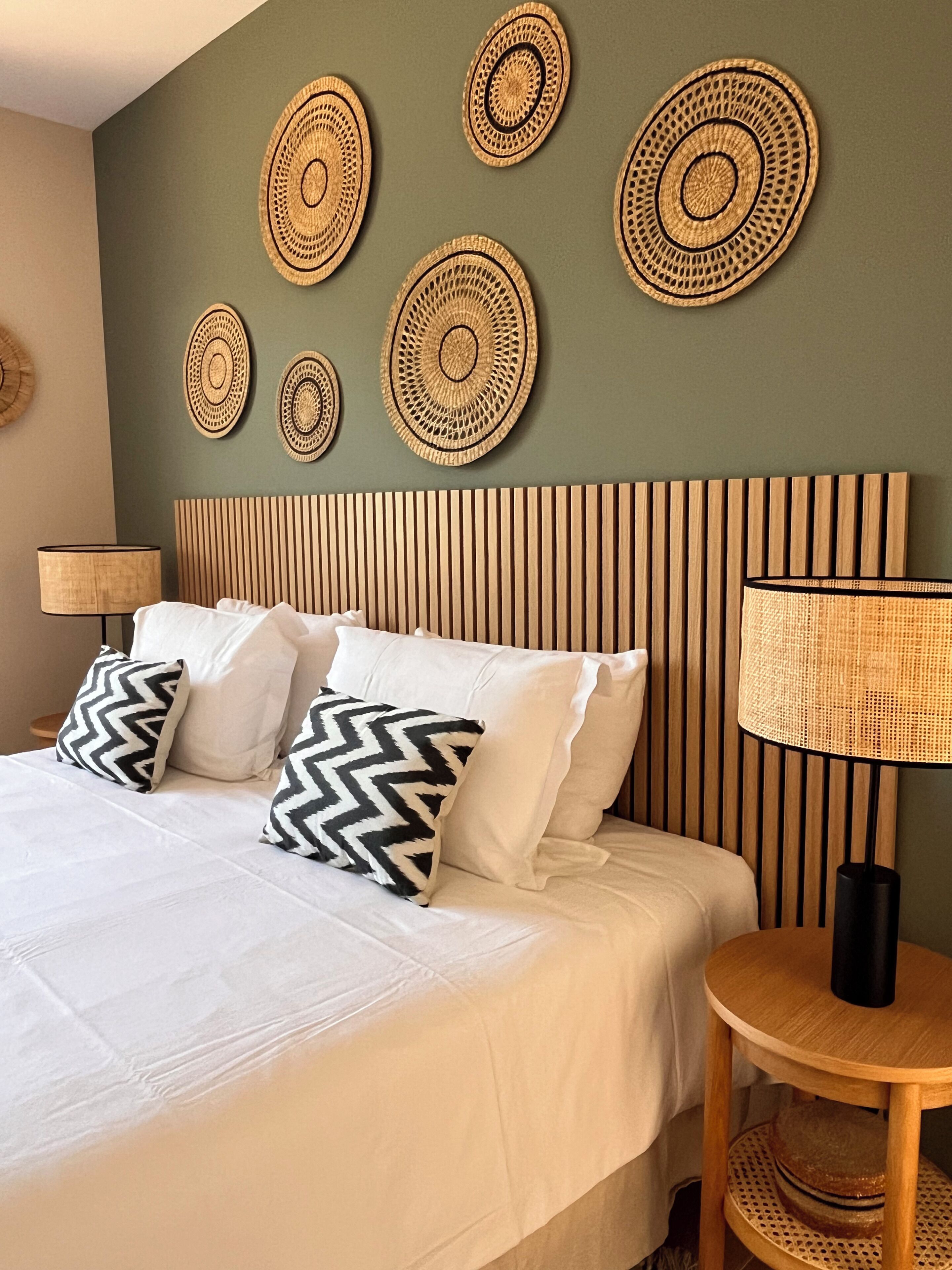 A bed with white bedding and patterned pillows, a wooden slatted headboard, and woven wall art.