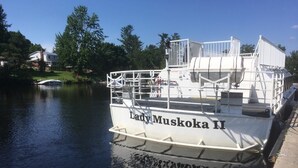 Un barco blanco llamado 'Lady Muskoka II' atracado en un muelle, con un cuerpo de agua tranquilo y vegetación al fondo.