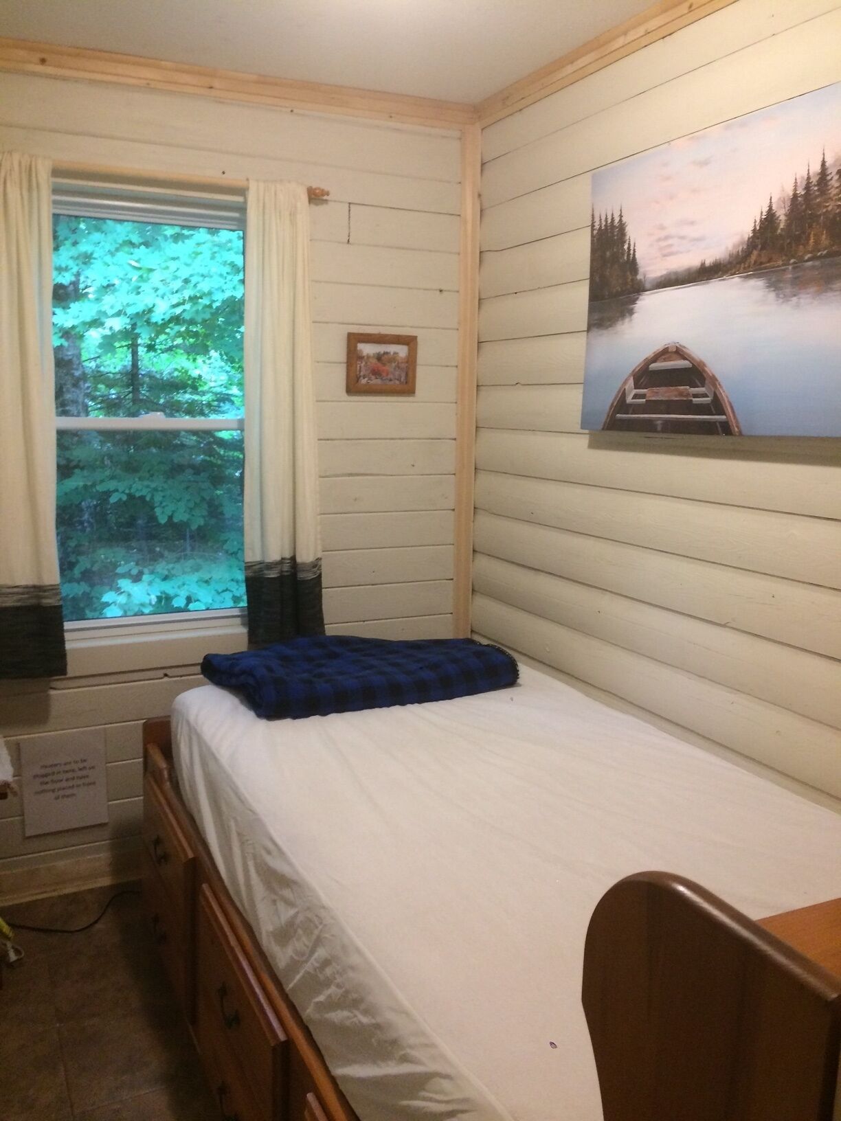 Ein kleiner, gemütlicher Raum mit einem Einzelbett, einem Fenster mit Vorhängen, einem Bild an der Wand und einem kleinen Bilderrahmen.