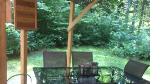 Una terraza exterior de madera con una mesa de vidrio y sillas de metal, rodeada de abundante vegetación.