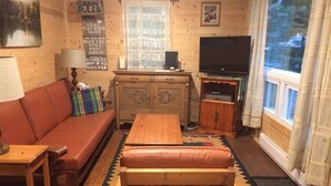 Una habitación tipo cabaña con sofá, un banco de madera, un mueble con televisor y una ventana con cortinas.