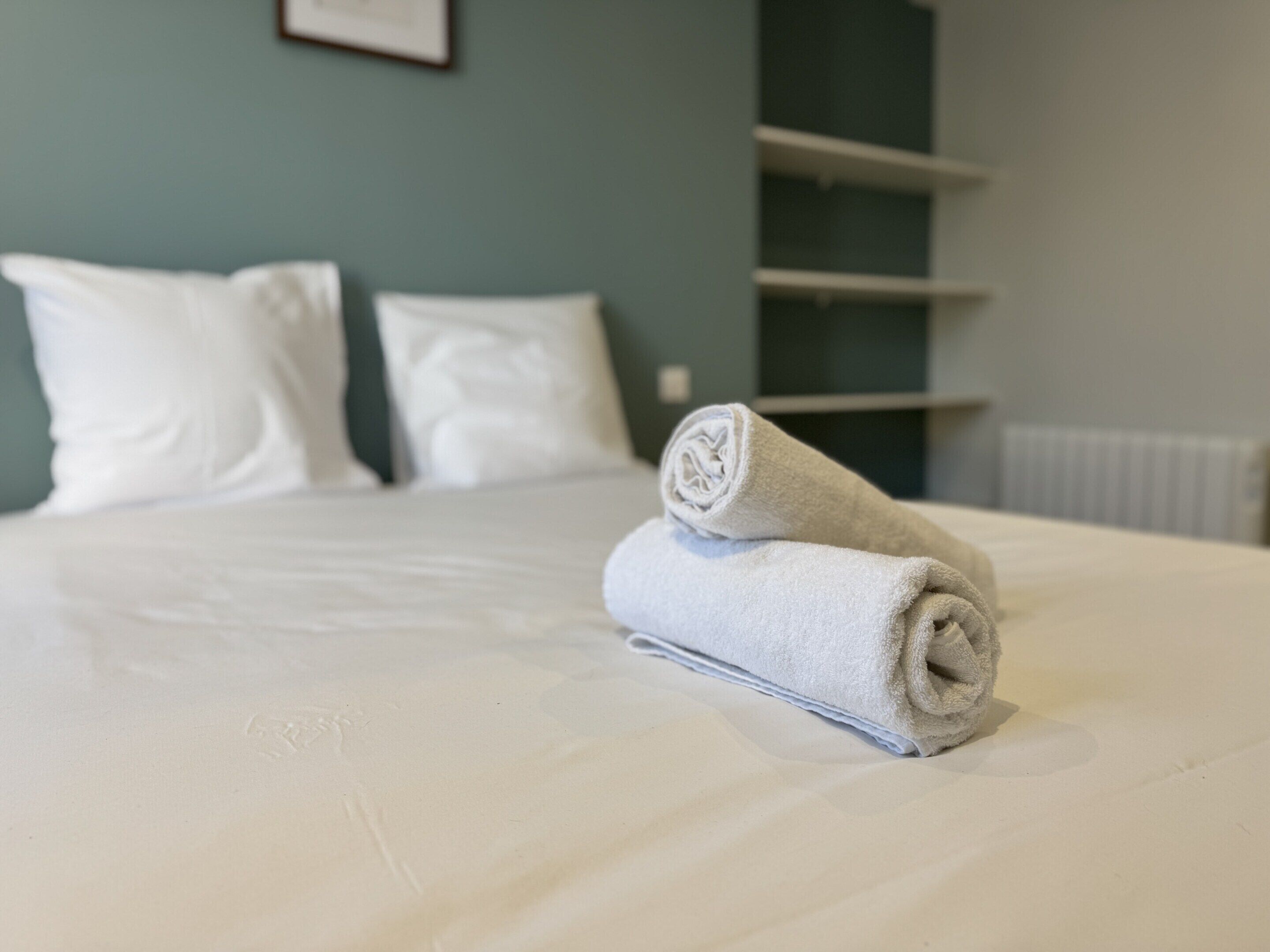 A neatly made bed with a rolled towel, white pillows, and a light blue wall with a framed picture.