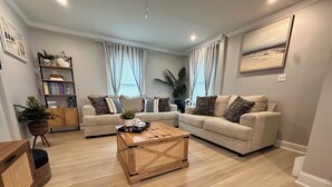 Un salon avec deux canapés beige, une table basse en bois, une bibliothèque et une grande fenêtre avec des rideaux.