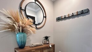 Une console en bois surmontée d’un miroir, un vase contenant des plantes séchées et une petite plante. Un étagère fixée au mur présente des objets décoratifs, et un sac est posé sur le sol.