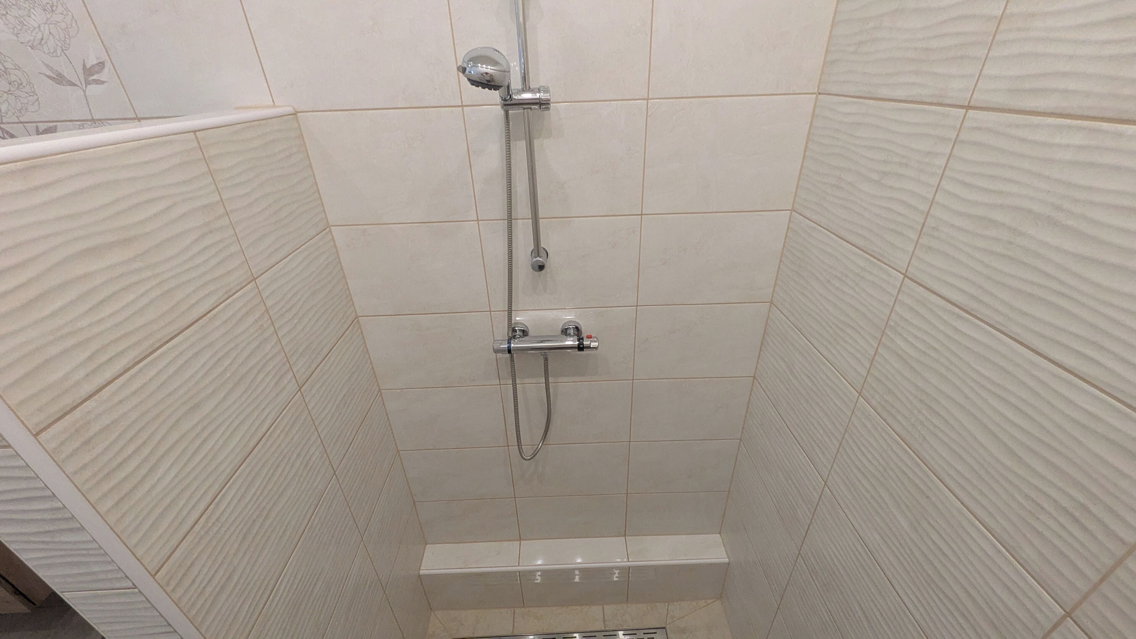 A shower with a handheld showerhead and a fixed showerhead, tiled walls, and a drain at the bottom.