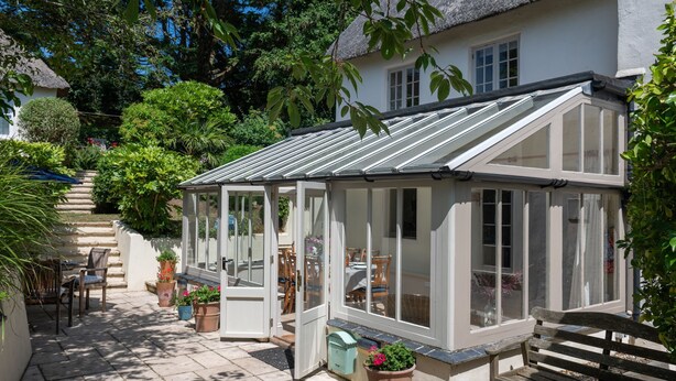 A conservatory with a glass roof, surrounded by potted plants and a wooden bench. - Thatchways -  a house that sleeps 7 guests  in 4 bedrooms (Thurlestone)