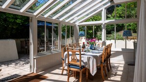 A conservatory with a glass roof, wooden chairs, a table set for dining, and a view of greenery outside. - Sea Lavender -  a house that sleeps 9 guests  in 5 bedrooms (Thurlestone)