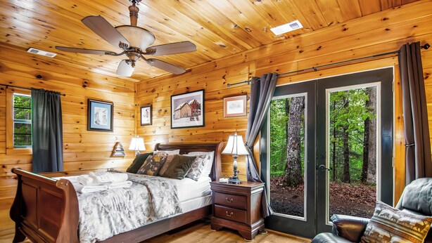 Une chambre à coucher dans un chalet en bois, avec un lit, des tables de chevet, une chaise et une grande fenêtre donnant sur la forêt.