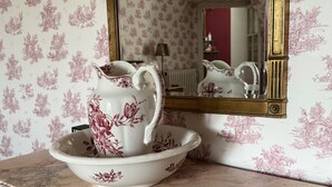 Ein Badezimmer mit einem мрамоrnen Waschtisch, einem weißen Krug mit floralen Verzierungen und einem Spiegel mit goldener Rahmung.
