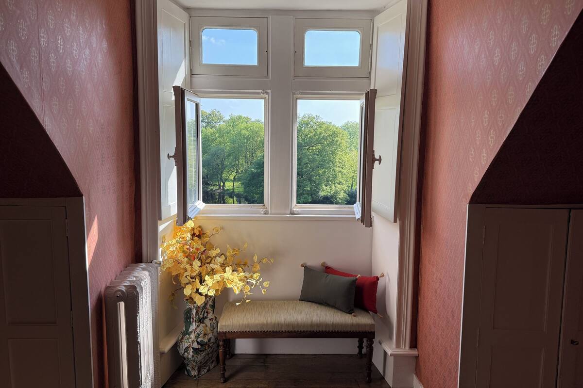 A room with a bench, a window with shutters, and a vase with yellow flowers.