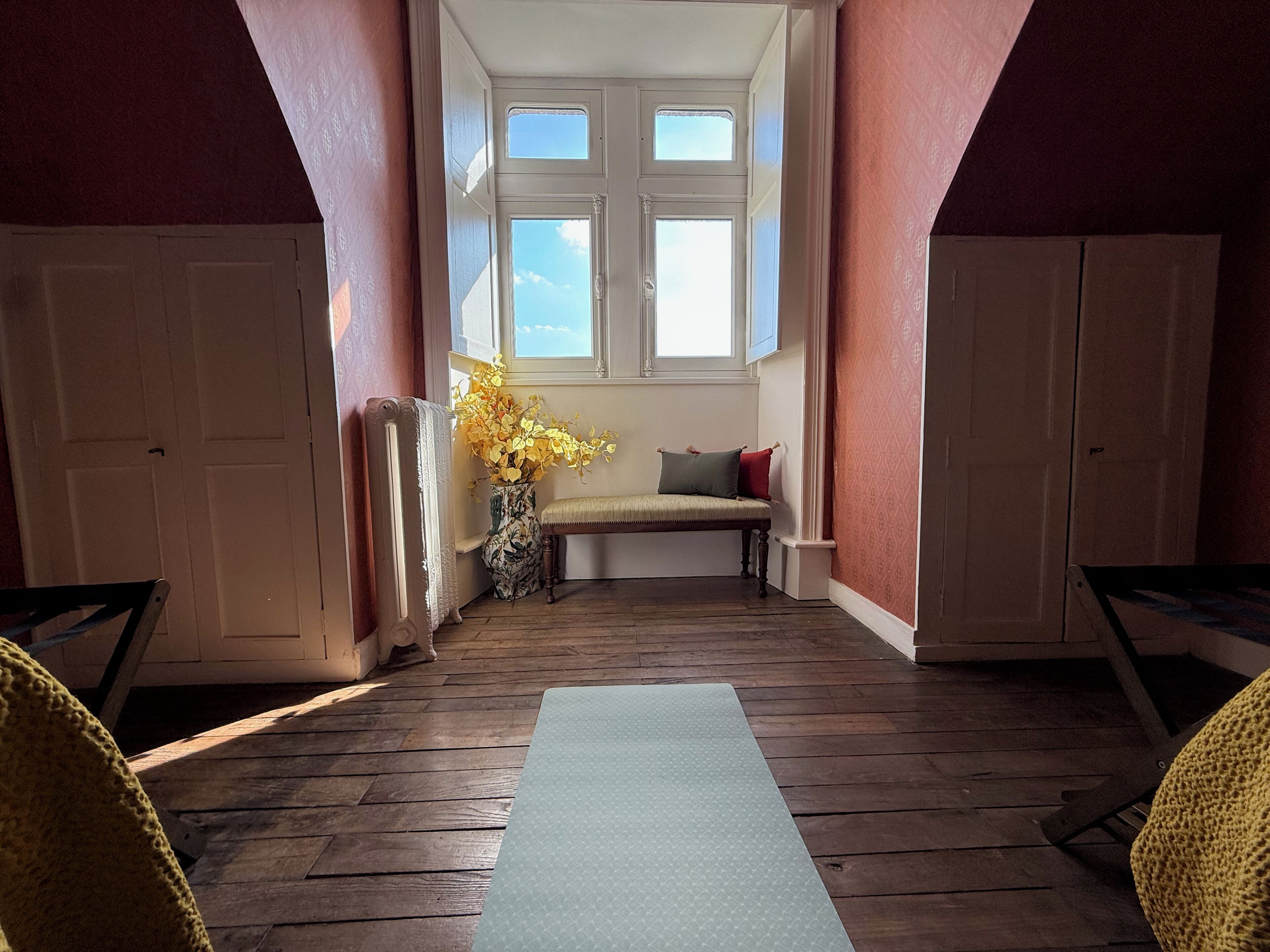A room with a wooden floor, a bench with cushions, a radiator, and a window with a view of the sky.