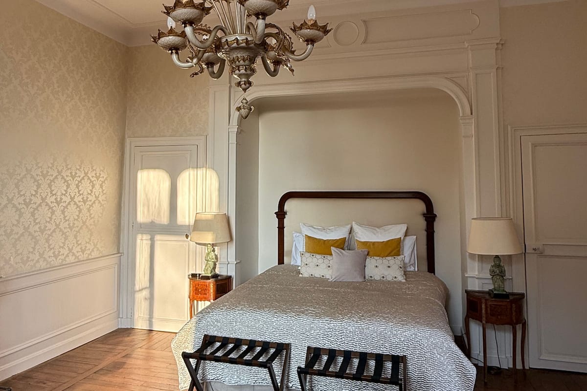 A bedroom with a large bed, a chandelier, a wooden bench, a patterned rug, and a nightstand with a lamp.