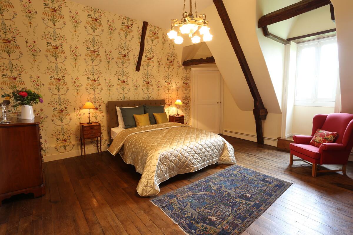 A bedroom with a bed, a red armchair, a wooden dresser, a nightstand, a lamp, and a patterned rug.