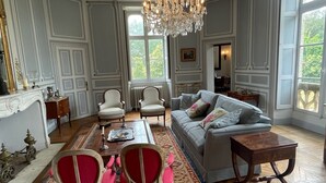 A spacious living room with a chandelier, a grey sofa, a red and white patterned rug, and a wooden herringbone floor. - Coin de Paix - Château de Launay (Saint-Vincent-sur-Oust)