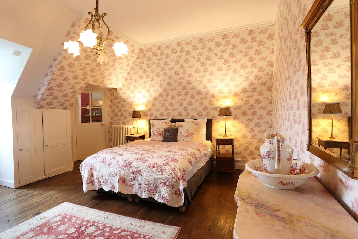 A bedroom with a sloped ceiling, floral wallpaper, a bed with a floral bedspread, bedside lamps, a dresser, a mirror, and a decorative bowl on the dresser.
