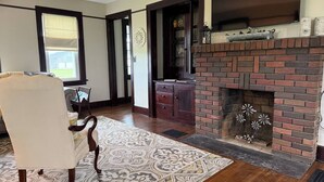A living room with a fireplace, wooden flooring, a patterned rug, and a dining area with a chair and table. - Charming 3-bedroom farmhouse with AC, WiFi in quaint Austinburg (Austinburg)