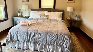 A neatly made bed with a quilted bedspread, two pillows, and a decorative pillow with the text "be our guest". There are two bedside tables with lamps and a small plant on one. The room has a large window with a view of a building outside. - Charming 3-bedroom farmhouse with AC, WiFi in quaint Austinburg (Austinburg)