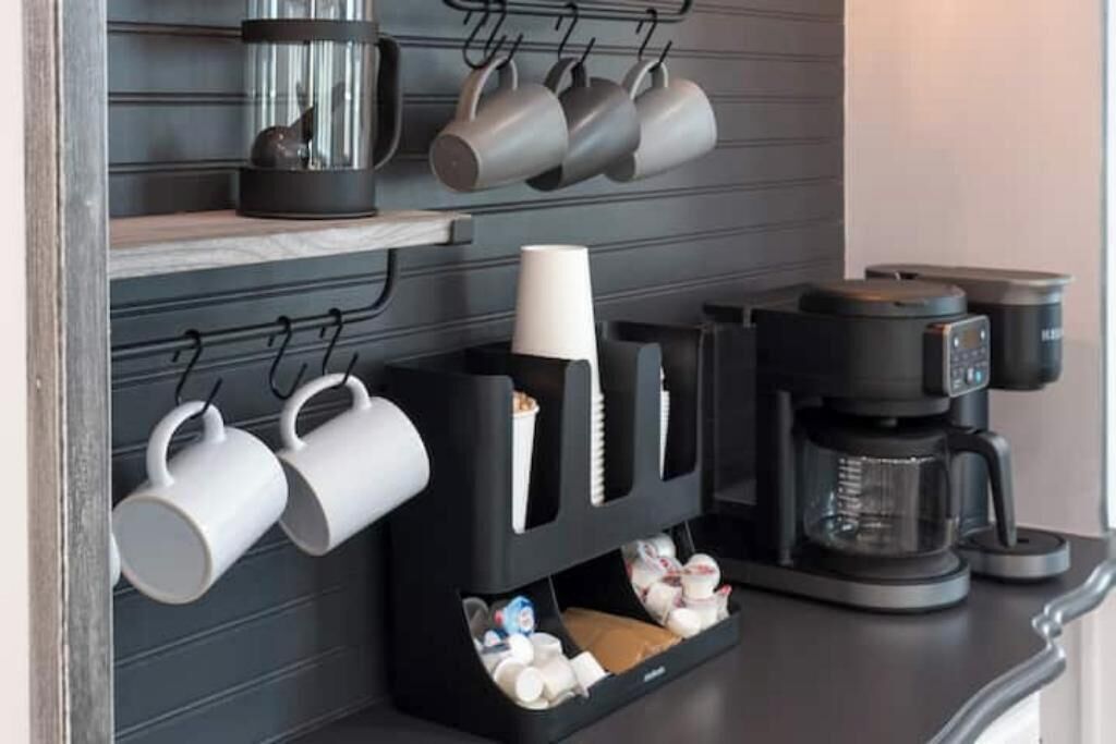 Une cuisine équipée d’une machine à café, de tasses suspendues à des crochets et d’un bac de rangement contenant des ustensiles.