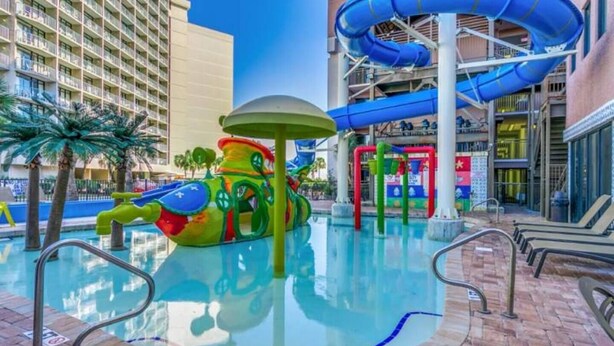A pool area with a water slide, a colorful play structure, and a building with balconies in the background.