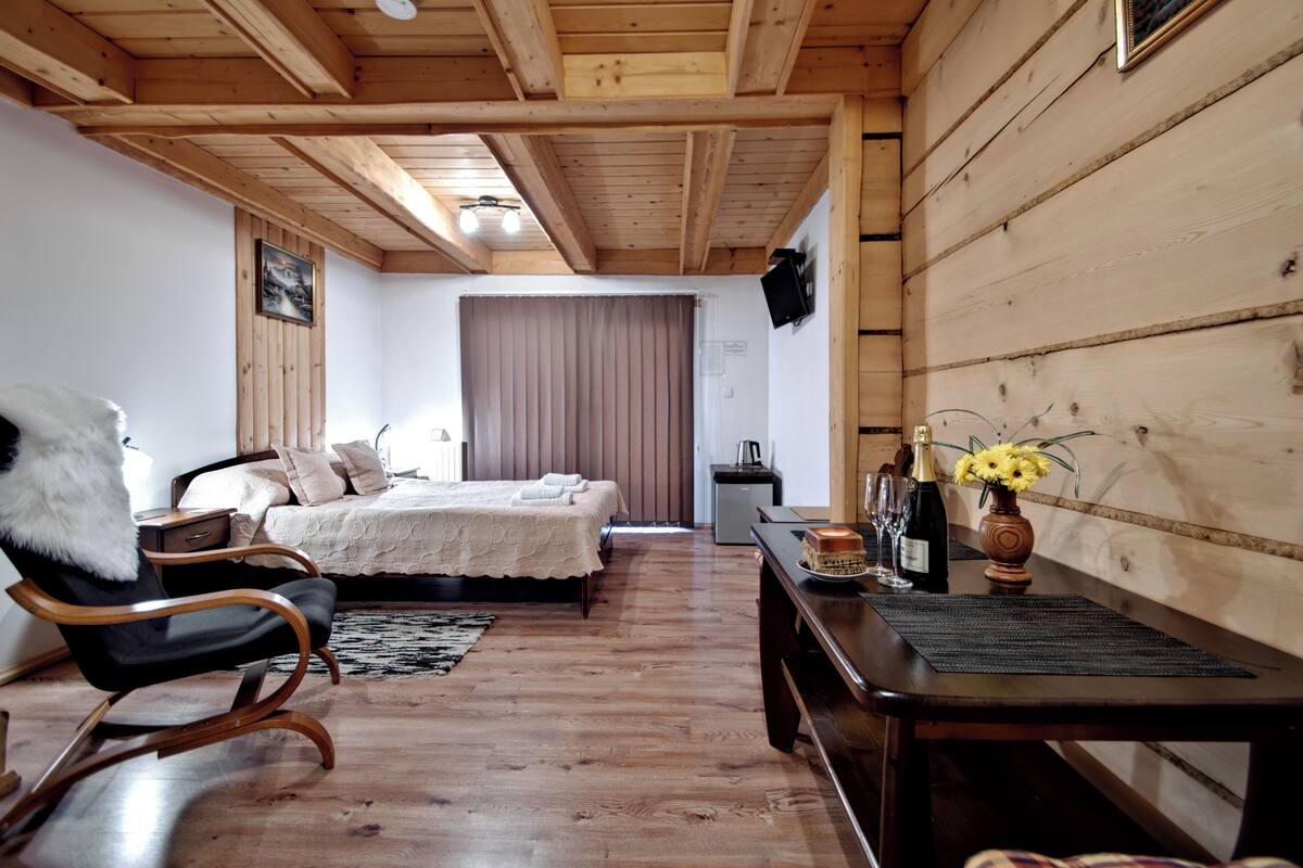 Una habitación con paneles de madera, una cama, una silla, una mesa con un jarrón y botellas, y un televisor en la pared.