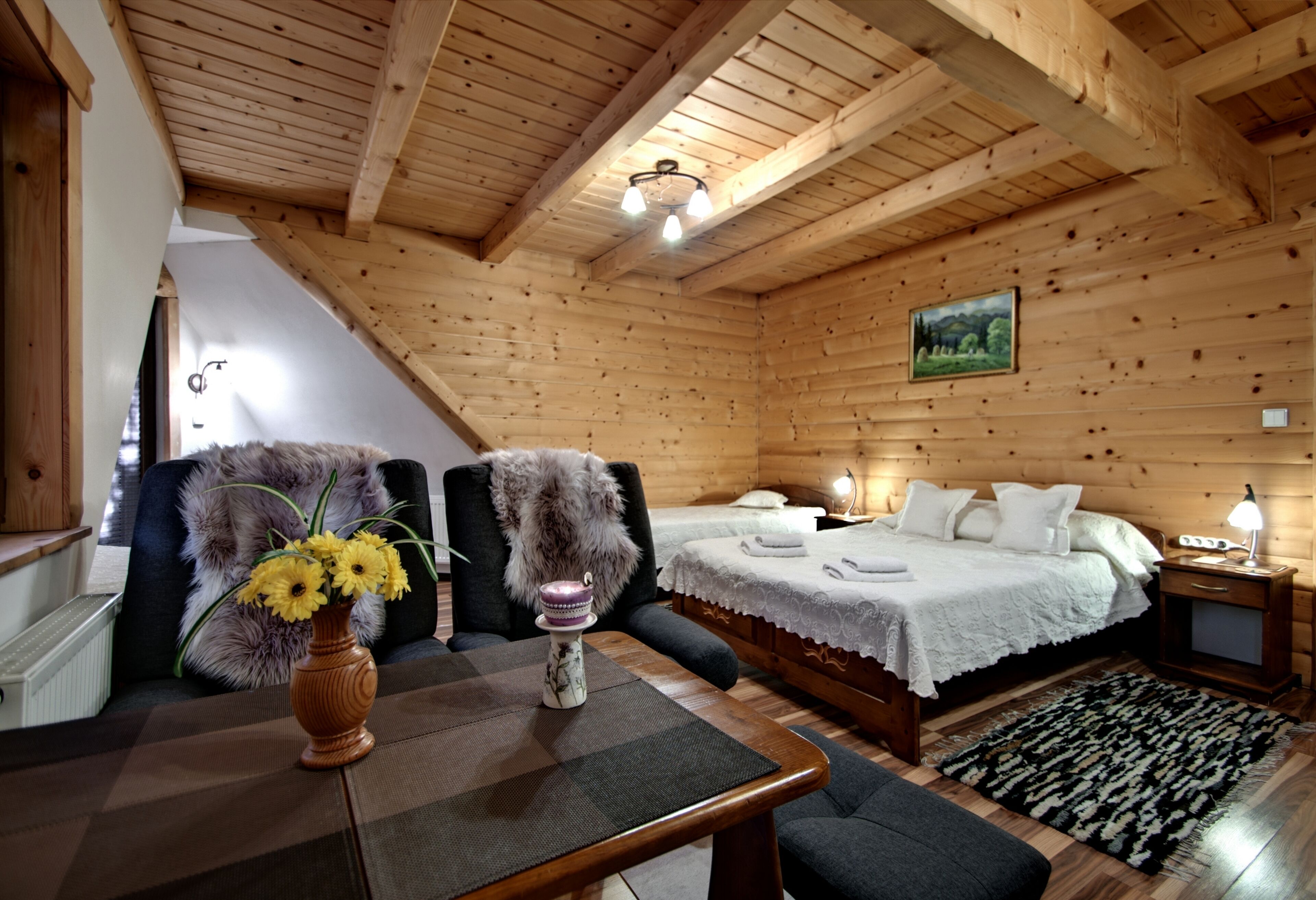 A wooden cabin interior with a bed, a table with a vase of flowers, a chair, and a painting on the wall.
