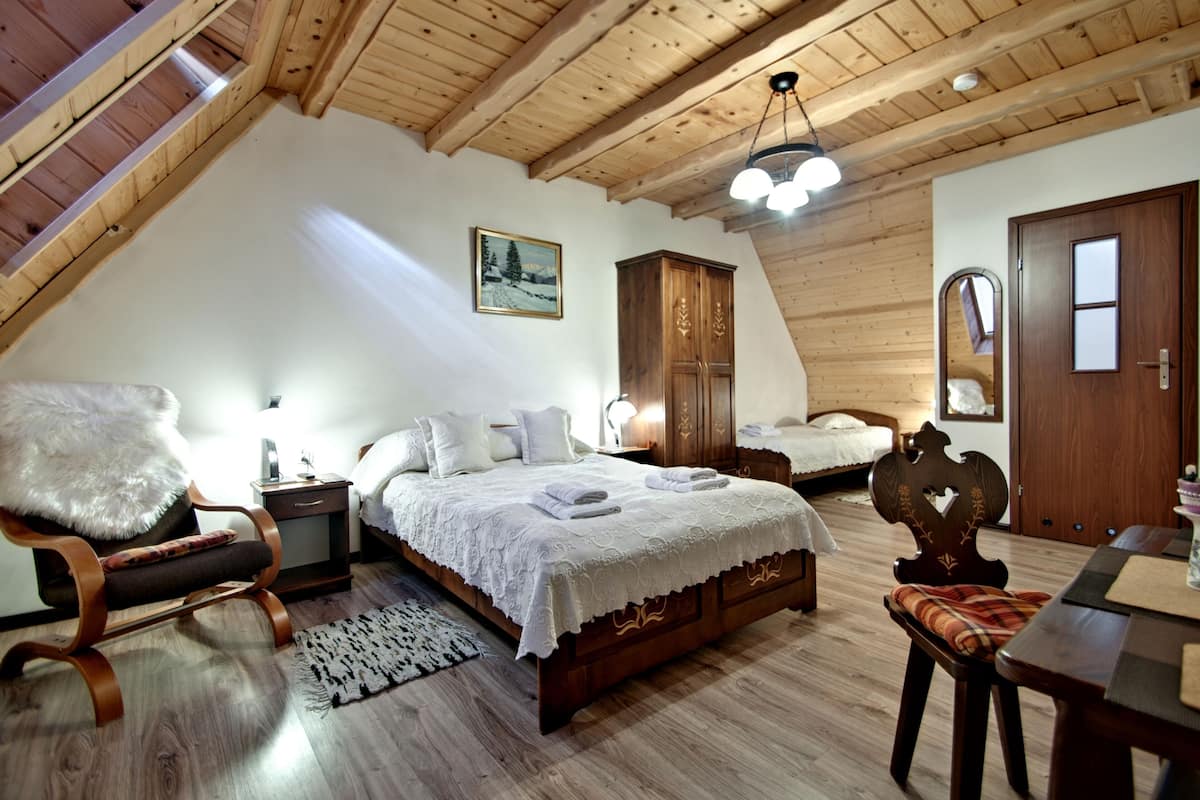 Un dormitorio tipo loft con techo inclinado, vigas de madera, una cama grande con ropa de cama blanca, un armario de madera, una silla y una mesa de comedor.