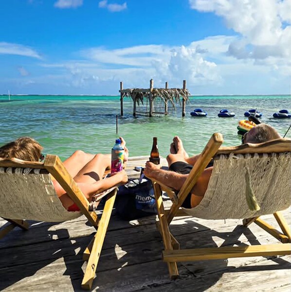 Two people relaxing on a wooden deck with a view of the ocean and a thatched structure in the background.
