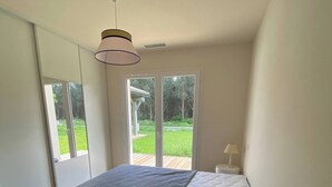 Ein Schlafzimmer mit einem Bett, einem weißen Nachttisch, einer Lampe und Blick auf eine grüne Außenanlage durch eine Schiebeglastür.