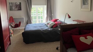 A bedroom with a bed, a desk, a chair, a radiator, a window with curtains, and a wall-mounted lamp. - Charming gîte in a hamlet near the Rance, between Dinard, Dinan and St Malo (Le Minihic-sur-Rance)