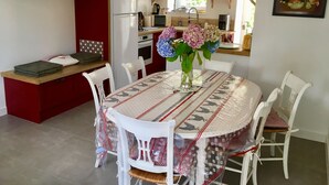 A dining area with a round table, white chairs, and a red kitchen in the background. - Charming gîte in a hamlet near the Rance, between Dinard, Dinan and St Malo (Le Minihic-sur-Rance)