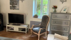 A living room with a flat-screen TV, a chair, a dresser, and a window with a view of greenery. - Charming gîte in a hamlet near the Rance, between Dinard, Dinan and St Malo (Le Minihic-sur-Rance)