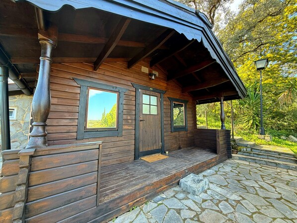 A wooden cabin with a stone-paved entrance, a covered porch, and windows overlooking a forested area.