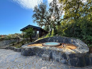 A stone hot tub surrounded by a stone wall, a wooden deck, and a building with a dark roof.