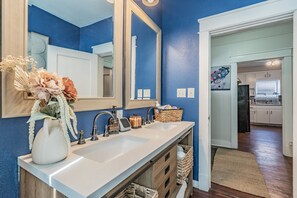 A bathroom with a white sink, a large mirror, and a vase of flowers. There is a basket of toiletries on the counter.