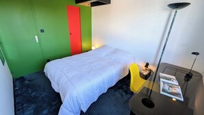 A small, modern room with a single bed, a green door, a red panel, and a small table with a lamp and a book.