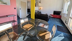 A modern living room with a glass dining table, black leather sofa, and a yellow accent wall.