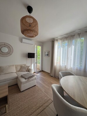 Un salon comprenant un canapé, une petite table, un fauteuil, un luminaire en fibres tressées, un téléviseur à écran plat et une fenêtre avec des rideaux.