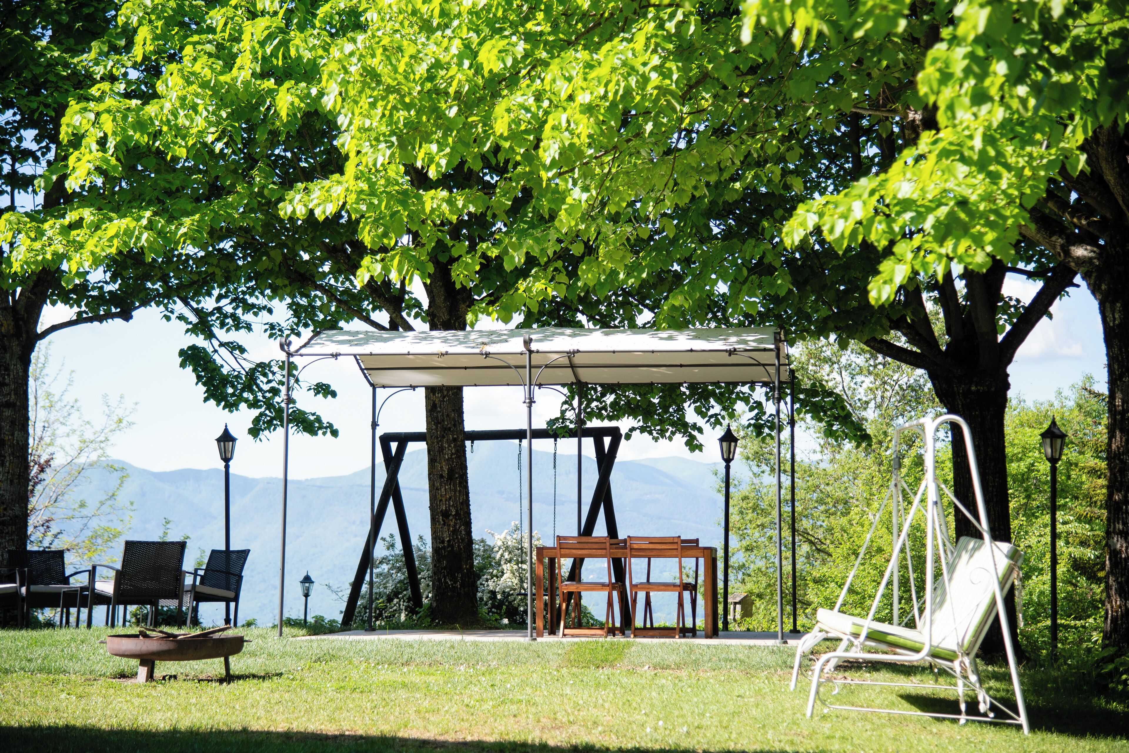 A gazebo with a swing chair and a table, surrounded by chairs and a boat, set against a backdrop of trees and mountains.