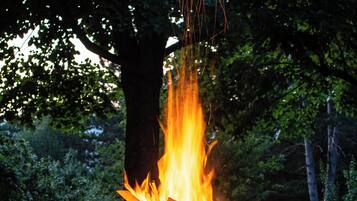 Een kampvuur in een metalen vuurkorf, omringd door gras, met bomen op de achtergrond.