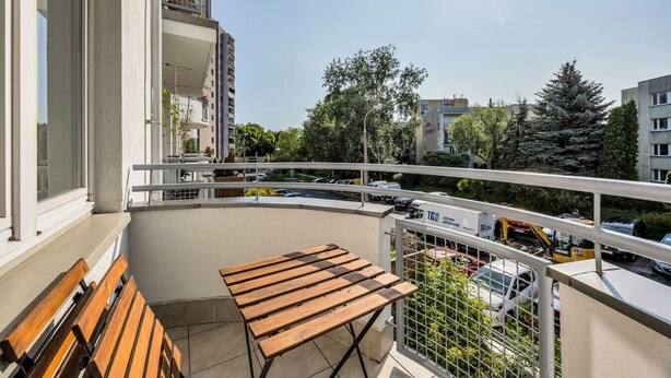 Varanda com banco e mesa de madeira, com vista para um parque de estacionamento e edifícios.