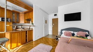 Um espaço compacto com cozinha compacta, cama com manta rosa e televisão de tela plana na parede.
